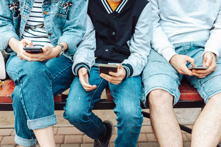 Attractive people family of different ages use a smartphone while while sitting together on a bench on the street. Woman and son in casual denim clothesの写真素材