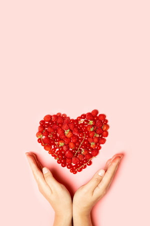 Beautiful fruit heart made of berries with female hands on a pink background with copy space. View from above. Vertical photoの写真素材
