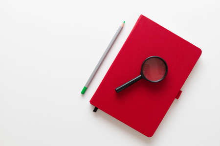 Home office desk table with red notebook, plant, magnifier and pencils on a white minimalistic background with copy space. Minimal work spaceの写真素材
