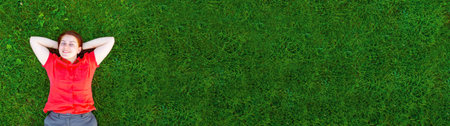Portrait of a smiling red-haired young girl on green grass looks at the camera outside in park....