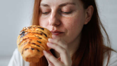 Happy young woman eats a croissant sitting on couch at home at morningの写真素材