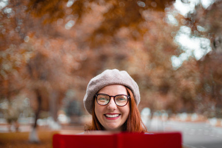 Young surprised girl in warm sweater and glasses holds a red notebook in the autumn park