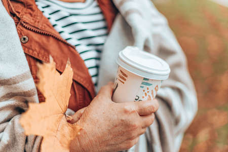 Caucasian woman holding hand with autumn maple leaf and paper cup with hot drink. Lady in warm blanket in the fall seasonの写真素材