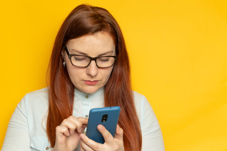 Focused business woman uses smartphone while standing against yellow wall background