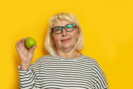Cheerful old blonde haired woman hold a green apple on a yellow background