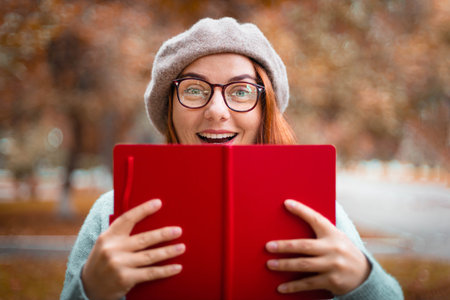 A young surprised girl in a beret and a warm sweater holds a red notebook in the autumn park