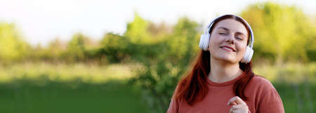 Beautiful young girl with eyes closed listening music through headphone and dancing on sunny day in city parkの写真素材