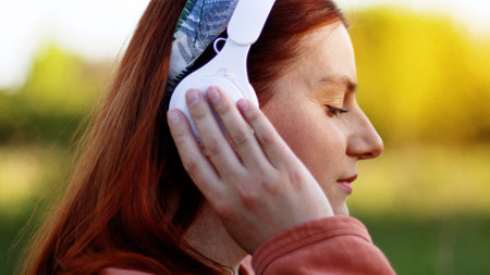 Attractive smart young woman listening music with headphones on sunny day while walking in city parkの写真素材