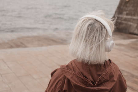 Back view of blonde woman wearing headphones listening music or podcast from smartphone application against the seaの写真素材