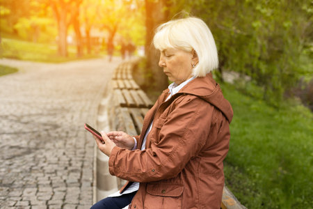 Active senior woman uses smartphone while sitting on a bench in the park