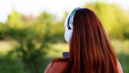 Back view of caucasian woman listening music with headphones on sunny day while walking in city parkの写真素材