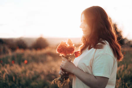 Happy young red-haired woman with flower in a blooming poppy field in the sun.の写真素材