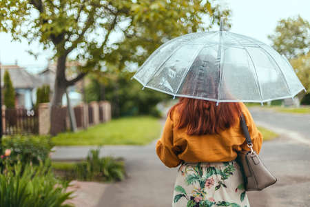 Back view of young woman in bright clothes under a transparent umbrella in bad weather in the city parkの写真素材