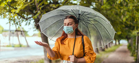 Unhappy beautiful woman in a protective medical mask standing under transparent umbrella and stretching hand catching water drops with contentの写真素材