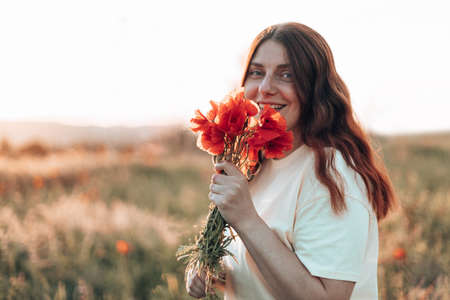 Happy young girl with a poppy bouquet in her hands in a summer field at sunset. Woman looking at the cameraの写真素材