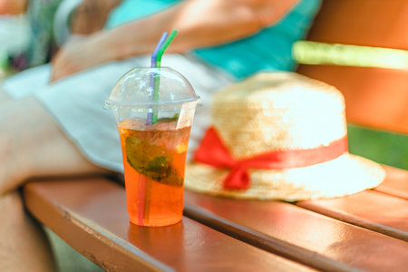 Close up takeaway plastic cup with aperol spritz cocktail on wooden bench in morning garden. Sunny day. Nature background.の写真素材