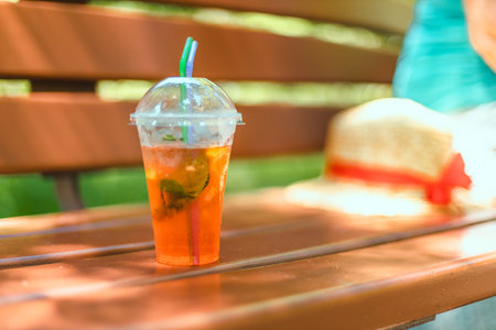 Close up takeaway plastic cup with aperol spritz cocktail on wooden bench in morning garden. Sunny day. Nature background.の写真素材