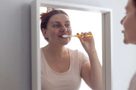 Portrait of beautiful millennial woman brushing teeth looking in mirror during morning hygiene procedures in the bathroom. Hygiene, dental care concept.の写真素材