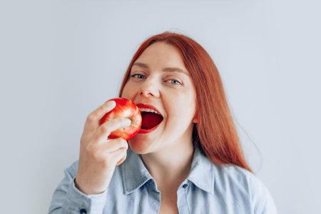 Portrait of a cheerful young woman eating red apple on gray wall background. Healthy nutrition diet....