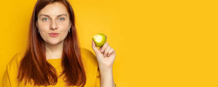 Proper nutrition balance concept. Young girl eats a green apple and smiles while standing against a yellow wall background. Proper nutrition concept, dietの写真素材