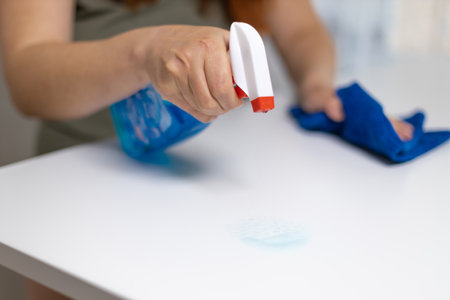 Close up of woman cleaning white table, sanitizing table surface with disinfectant antibacterial spray bottle, washing surfaces with towel and gloves. COVID-19 prevention sanitizing insideの写真素材