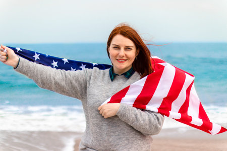 Caucasian woman holding american flag leaning against back and looking at a sea. Usa flag for...