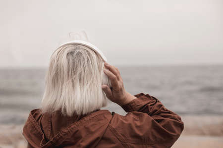 Back view of blonde woman wearing headphones listening music or podcast from smartphone application against the seaの写真素材