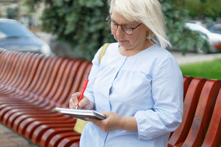 Mature businesswoman writing info thoughts on notepad outdoors in a city park in summer outdoors, education conceptの写真素材