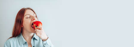 Cheerful young woman eating red apple on gray wall background. Healthy nutrition diet. Apple vitamin snack.の写真素材