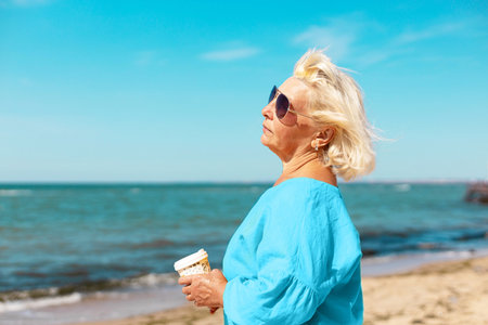 An adult woman in sunglasses and a yellow blouse with a paper cup of coffee to go by the sea on a...