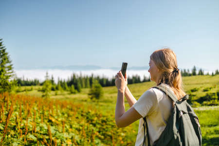 Travel female photographer with backpack and smart phone standing on woody mountains landscape and bright sun in blue sky background. Amazing Carpathians landscape backgroundの写真素材