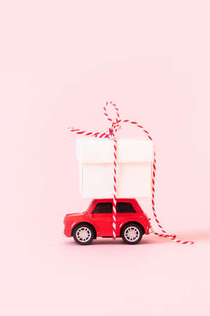 Christmas and New Year holiday background. Bright red model car toy with a surprise gift on the hood on a pink background. Vertical photoの写真素材