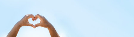 Female hands making a heart gesture in front of blue skyの写真素材