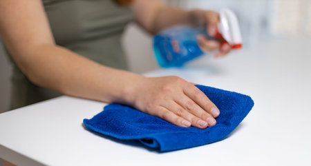 Close up of woman cleaning white table, sanitizing table surface with disinfectant antibacterial...