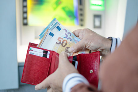 Unrecognizable female puts out a 50 and 20 euro banknote from a red leather wallet near the ATM on the street.の写真素材