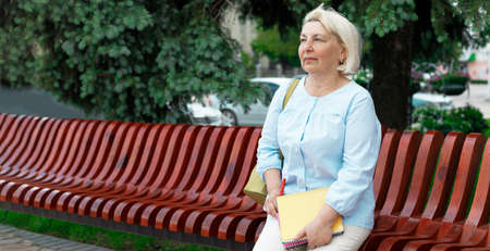 Adult 50 years old teacher woman on a wooden bench in a park holding booksの写真素材