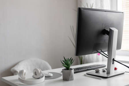 Modern computer monitor is located in a work place in a bright interior. Stylish white wireless headphones and a flower on a white table.の写真素材