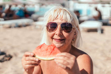 Adult blond caucasian woman in sunglasses eating big piece of watermelon sitting on the beach in summer near the seaの写真素材