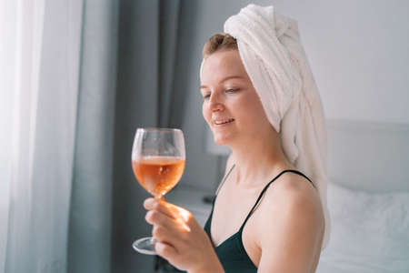 Morning wine in bed. Young european woman sitting in bed with wine glass in cozy comfort bed after waking up. Alcohol, celebration and mourning.の写真素材