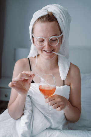 Happy woman with towel on head lies in bed under blanket isolated over white wall background drinking wine, have a fan Alcohol, celebration and mourning.の写真素材