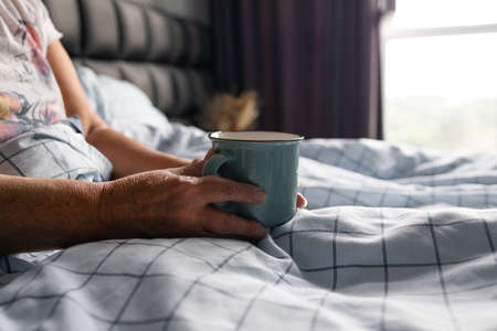Crop photo of relaxing woman enjoying sunny morning holding cup in bedの写真素材