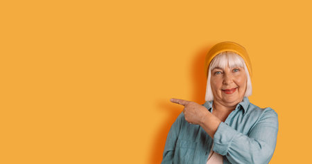 Happy 50s blonde woman in stylish modern clothes posing against a yellow background, showing free...