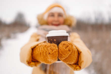 Happy woman hands in yellow knitted mittens holding hot chocolate or coffee for taking away on urban winter streetの写真素材