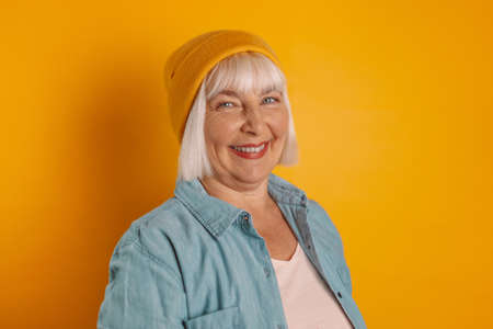 Photo of optimistic older blond lady in a yellow hat and stylish casual clothes smiling at the camera isolated over color background, People lifestyle conceptの写真素材