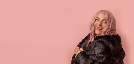 Portrait of happy and positive caucasian woman with pink curly hair wears black winter fur coat enjoying free time isolated over pink background. Happiness conceptの写真素材