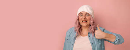 Attention promotions banner. Charming young caucasian woman in a white knitted hat with pink hair showing thumb up like gesture isolated on pastel background, banner, panorama, copy spaceの写真素材