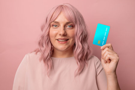 Attractive smiling young woman holds credit card, thinking about shopping isolated on pink studio...