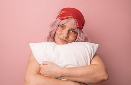 Sweet attractive lovely woman wear sleep mask, hugging pillow looking at the camera isolated over...