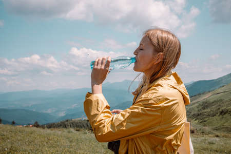 Tired traveler caucasian woman hiker in a yellow waterproof jacket drinking water from bottle while climbing on mountain in Carpathians, Ukraineの写真素材