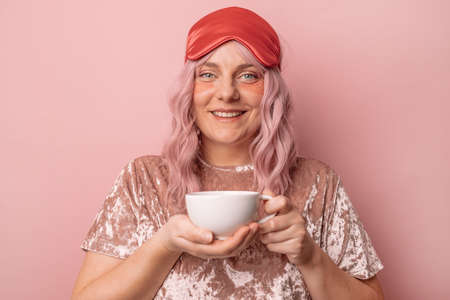 Happy smiling attractive woman wears a sleep mask holding a cup of fragrant coffee or tea in the morning isolated over pink background.の写真素材